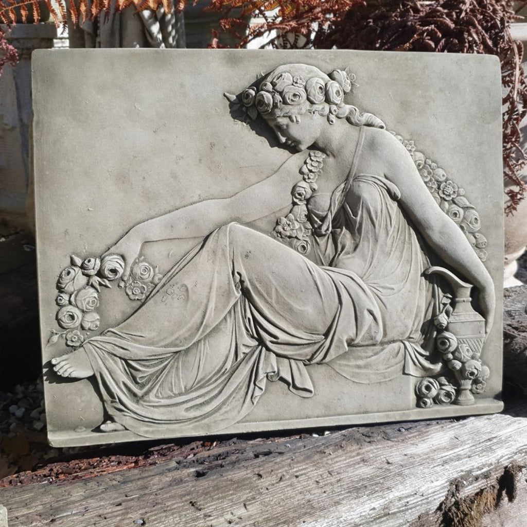 A rectangular stone garden plaque featuring a classical relief sculpture of a draped female figure with flowers, displayed outdoors on a weathered wooden surface.