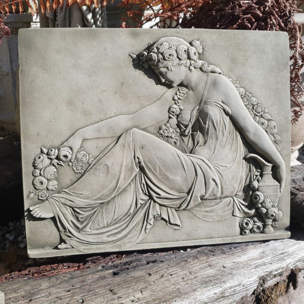 A rectangular stone garden plaque featuring a classical relief sculpture of a draped female figure with flowers, displayed outdoors on a weathered wooden surface.