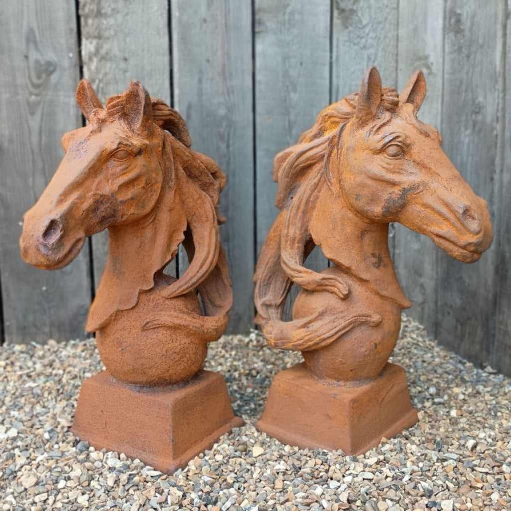 Two rusted metal horse head bust sculptures on square bases, displayed side by side on gravel in front of a weathered wooden fence.